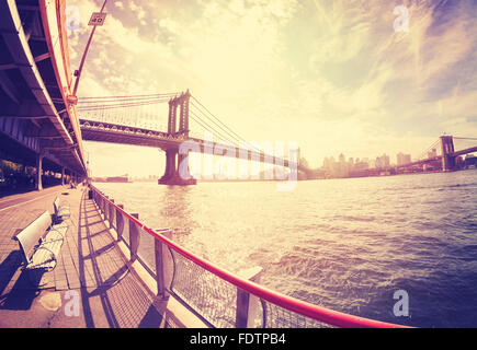 Vintage stylized fisheye lens photo of bench at Hudson River bank and Manhattan Bridge in distance against sun, NYC, USA. Stock Photo