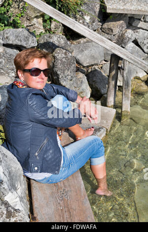 white middle aged red haired tanned woman in sunglasses, jeans and leather jacket sits barefoot on stones by the lake shore with Stock Photo