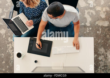 Two young people doing graphic design work. Man working on graphics tablet and computer while woman using laptop. Both sitting t Stock Photo