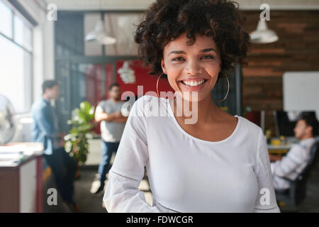 Portrait of smiling young african businesswoman with people in background. Cheerful young woman standing relaxed in her office, Stock Photo