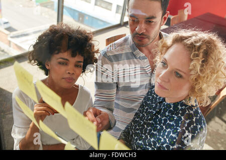 Three coworkers discussing new ideas on a glass wall. Creative professionals standing at the office behind glass wall with stick Stock Photo