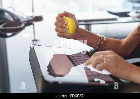 Office worker typing email on tablet computer. The woman feels stressed and nervous, holds an antistress yellow ball in her hand Stock Photo
