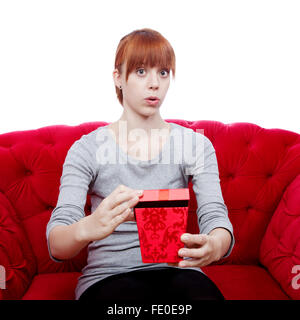 young beautiful red haired girl opens gift box for you on red sofa in front of white background Stock Photo