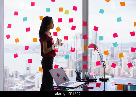 Mixed race secretary working in modern office in skyscraper, writing and sticking adhesive notes with tasks on window. Stock Photo