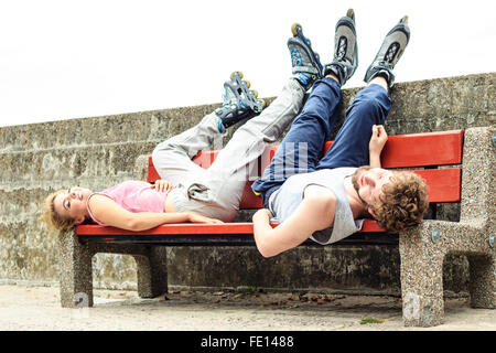 Young tired people friends in training suit with roller skates. Woman and man relaxing lying on bench outdoor. Stock Photo