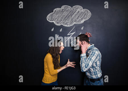 Crazy young couple standing under drawn raincloud and yelling on each other over blackboard background Stock Photo