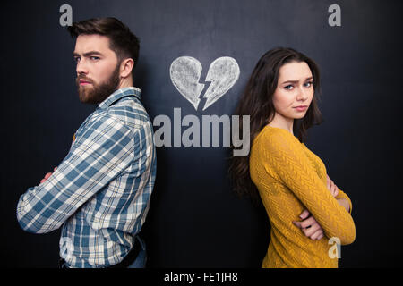 Unhappy young couple standing with arms crossed over background of chalkboard with drawn broken heart Stock Photo