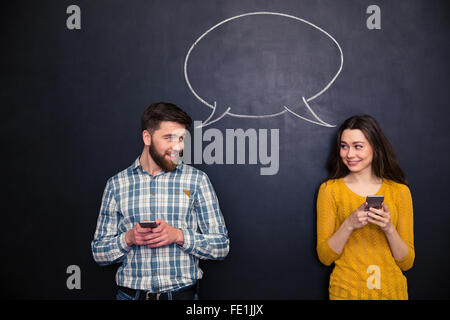 Beautiful couple using smartphones and flirting over blackboard background with drawn speech dialogue Stock Photo