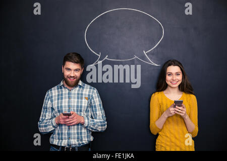 Happy young couple using mobile phones and smiling over chalkboard with drawn blank speech bubble Stock Photo