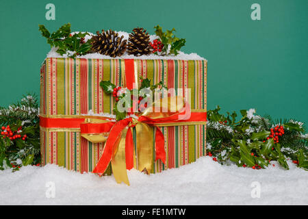 A gift wrapped Christmas present surrounded by a holly garland and snow, green background. Stock Photo