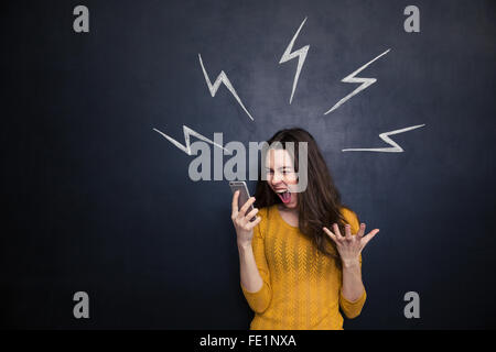 Crazy young woman using smartphone and yelling over black board with drawn lightnings behind her Stock Photo
