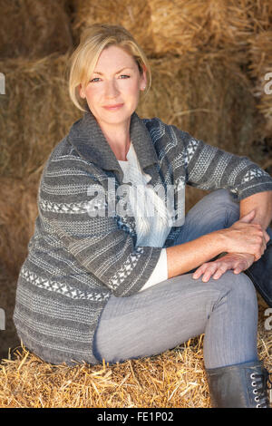 Attractive middle aged blond woman sitting on a hay bale in a barn wearing boots Stock Photo