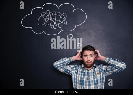 Confused bearded young man grabbing his head and thinking about problem with black board behind him Stock Photo