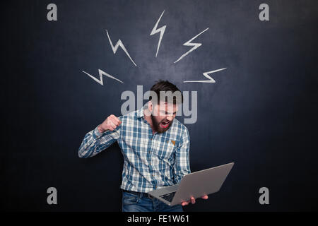 Aggressive young man yelling and going to break laptop with fist over chalkboard background Stock Photo