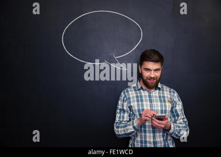 Smiling young man using smartphone over blackboard background with empty speech bubble Stock Photo