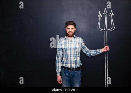 Smiling bearded young man standing and holding drawing trident over chalkboard background Stock Photo