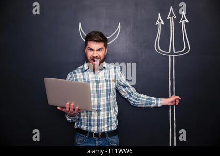 Angry bearded man using laptop and holding trident drawn on blackboard background Stock Photo