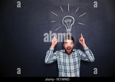 Surprised happy young man with beard pointing up with both hands over chalkboard background Stock Photo