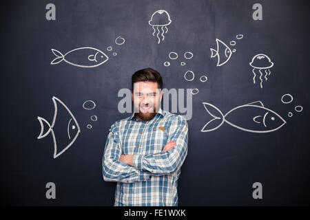 Cheerful handsome bearded young man standing with folded hands over blackboard background with drawn fishes and jellyfishes Stock Photo