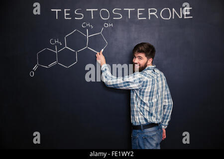 Happy bearded young man standing and drawing testosterone molecule chemical structure on chalkboard background Stock Photo