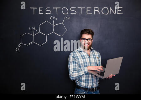 Happy young scientist standing over chemical structure of testosterone molecule drawn on chalkboard background and using laptop Stock Photo
