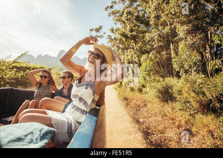 Portrait of a cheerful young woman sitting in the back of pickup truck with friends. Young people enjoying on a road trip. Stock Photo