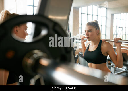 Young woman in gym doing squats with extra weight on shoulders. Fitness female working out in front of mirror at health club. Stock Photo