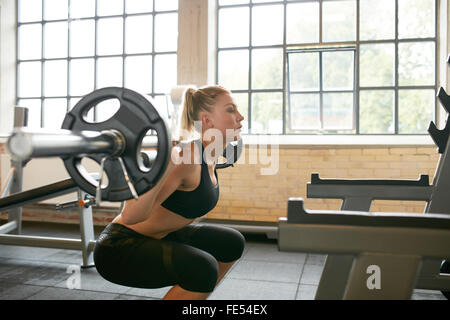 Female working out in a gym doing squats. Young woman working out using barbell with heavy weights in a fitness club. Stock Photo