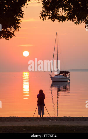 Waterfront scene of a Sailboat and photographer at sunrise, with a reflection in the water. Stock Photo