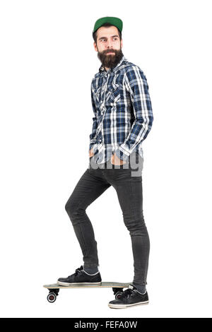 Happy confident hipster with hands in pocket wearing cap standing on longboard side view. Full body length portrait isolated Stock Photo