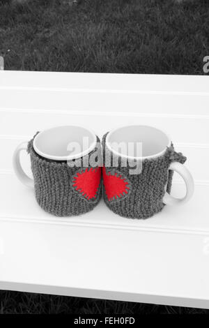 a vertical frontal black and white view of two mug with warmers creating the shape of a red heart in a white table on a garden w Stock Photo