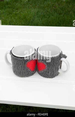 a vertical frontal view of two mug with warmers creating the shape of a heart in a white table on a garden with space for a mess Stock Photo
