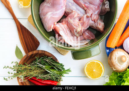 Top flat view of raw rabbit meat with vegetables in round ceramic pot on white wooden table surface Stock Photo