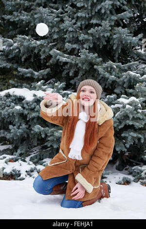 Girl play snowballs in winter forest at day. Snowy fir trees. Redhead woman full length. Stock Photo