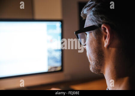 Concentrated bristled young man in glasses sitting and looking at screen of monitor Stock Photo