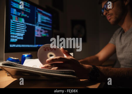 Confident young programmer taking cd with base and coding on computer in dark room Stock Photo