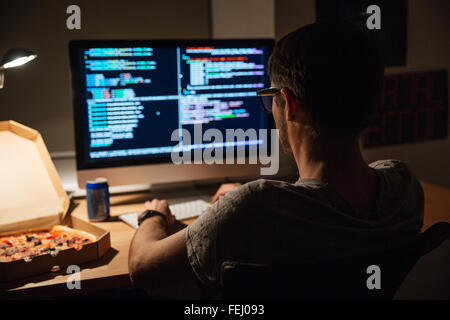 Back view of focused young programmer in glasses writing code and eating pizza Stock Photo