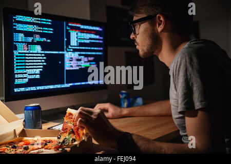 Profile of concentrated young software developer eating pizza and coding at home Stock Photo