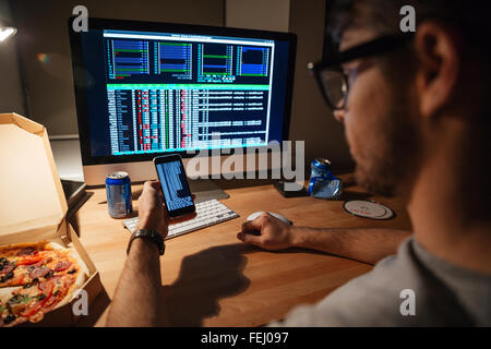 Closeup of serious young developer in glasses using computer and smartphone for coding at home Stock Photo