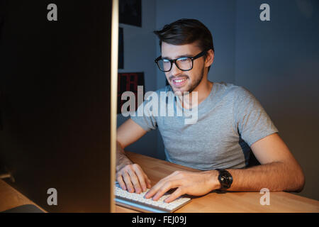 Happy attractive young man in glasses sitting and typing on computer at home Stock Photo
