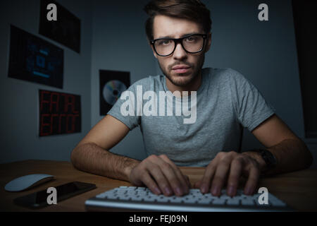 Pensive young man in glasses sitting at home and typing on keyboard Stock Photo