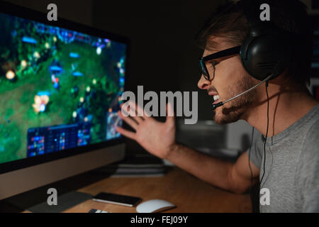 Cheerful young gamer in glasses playing game on computer using headphones Stock Photo