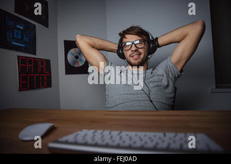 Relaxed carefree young man in glasses sitting with hands behind head and listening to music from computer Stock Photo