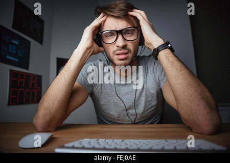 Frowning young man in earphones and glasses sitting with hands on head in front of computer Stock Photo