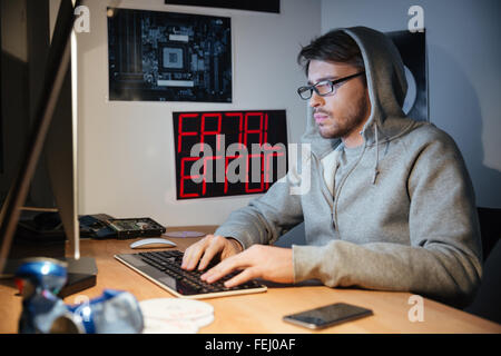 Handsome young man in sweatshirt with hood sitting and typing on computer keyboard at home Stock Photo