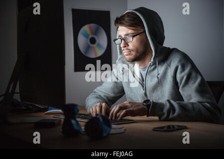 Thoughtful young man in glasses and hoodie working on computer in dark room at home Stock Photo