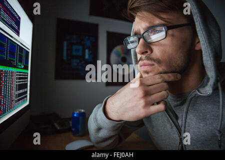 Pensive attractive young man in glasses and sweatshirt with hood thinking and coding at home Stock Photo