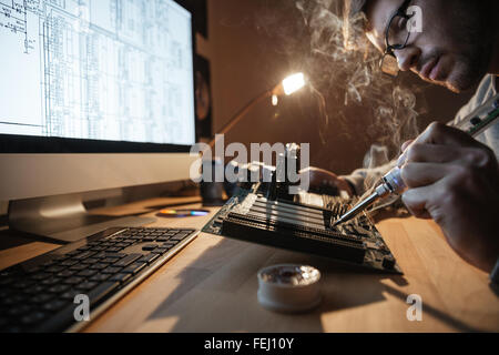 Concentrated young man in glasses looking at scheme on monitor and repairing motheboard with soldering iron Stock Photo