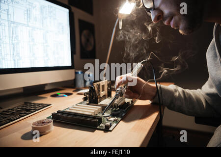 Closeup of serious young man using scheme on monitor and soldering iron for repairing motheboard Stock Photo