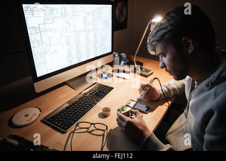 Serious handsome young man using soldering iron for fixing smartphone sitting in dark room Stock Photo